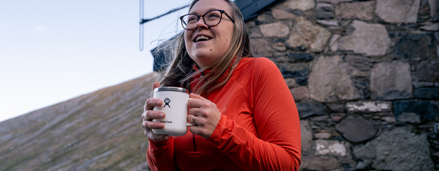 A woman, wearing a red baselayer, is holding a white mug outdoors 