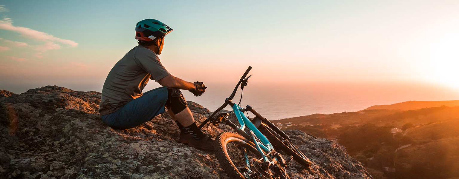 A person in full cycling kit is resting on a hill, with a bicycle next to him or her.