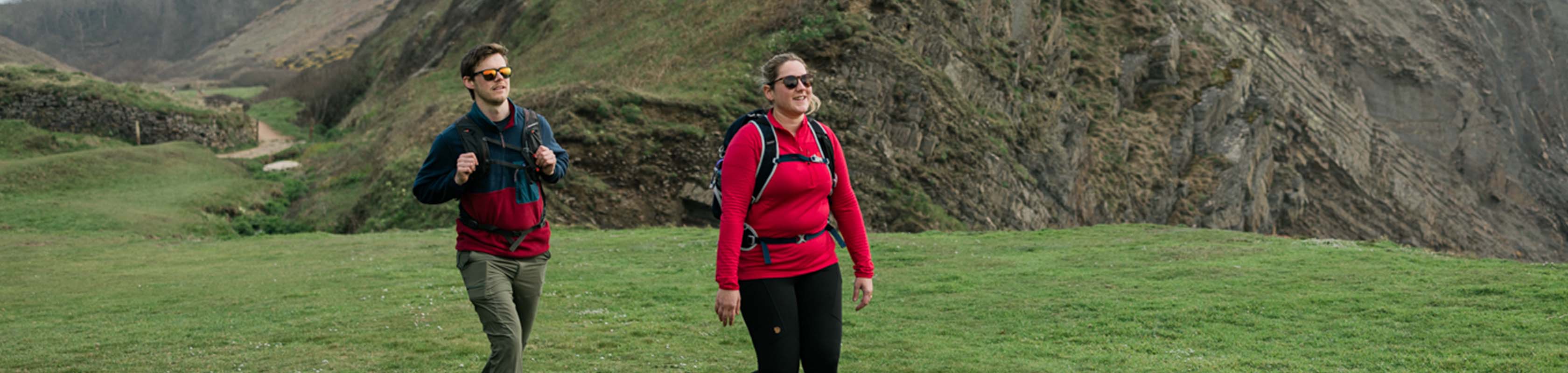 Two people are walking next to some hills, wearing fleeces, walking boots, and trousers, with rucksacks.