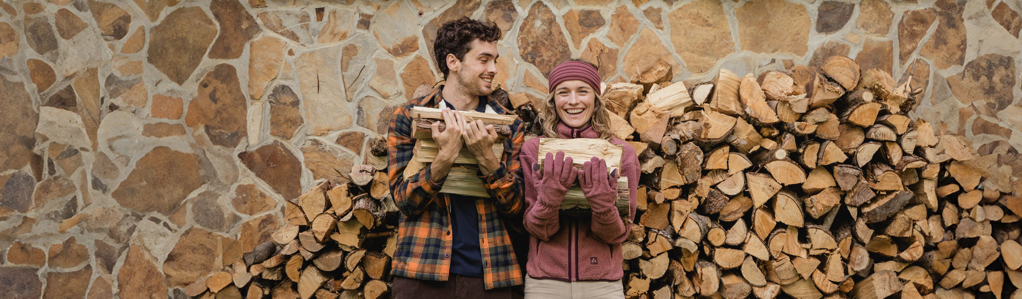 Two people stood holding logs, stood in front of a stack of logs, wearing their Ayacucho clothing 