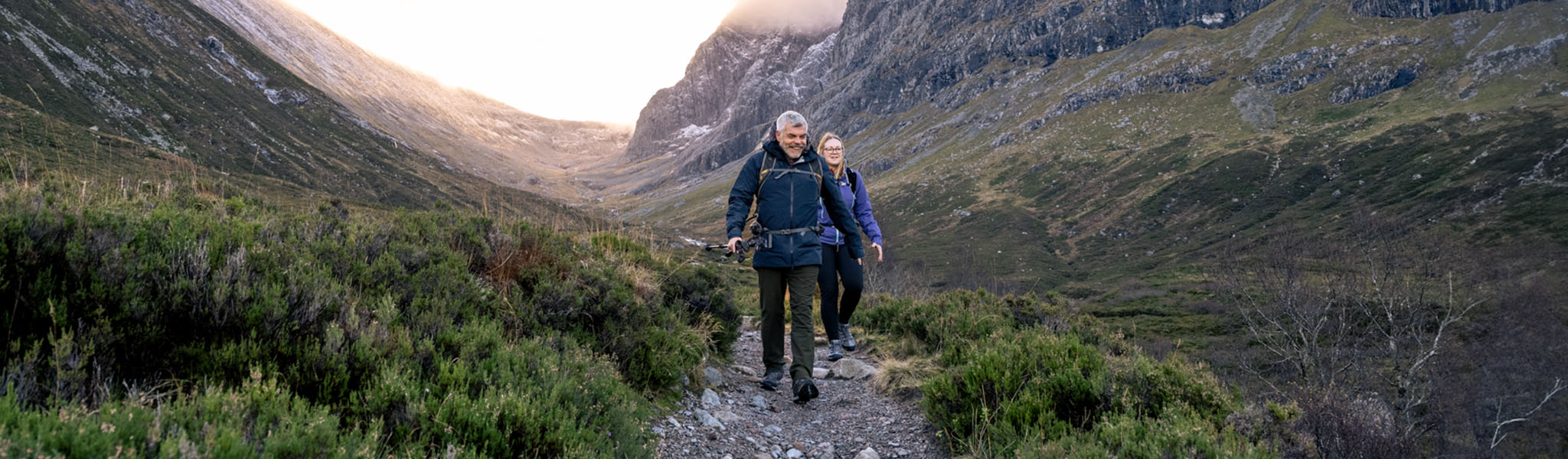 Two people are walkingon a path amongst hills and mountains.