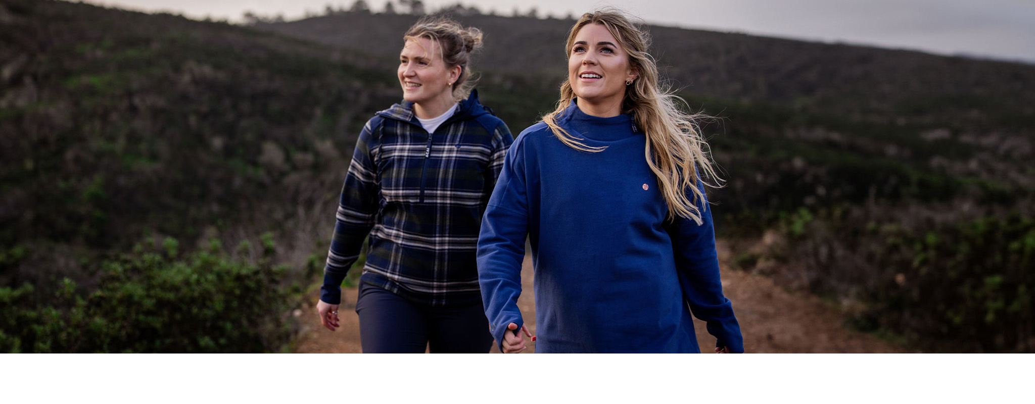 Two girls with blonde hair walk towards the camera with the hills behind in a blue and plaid jumper