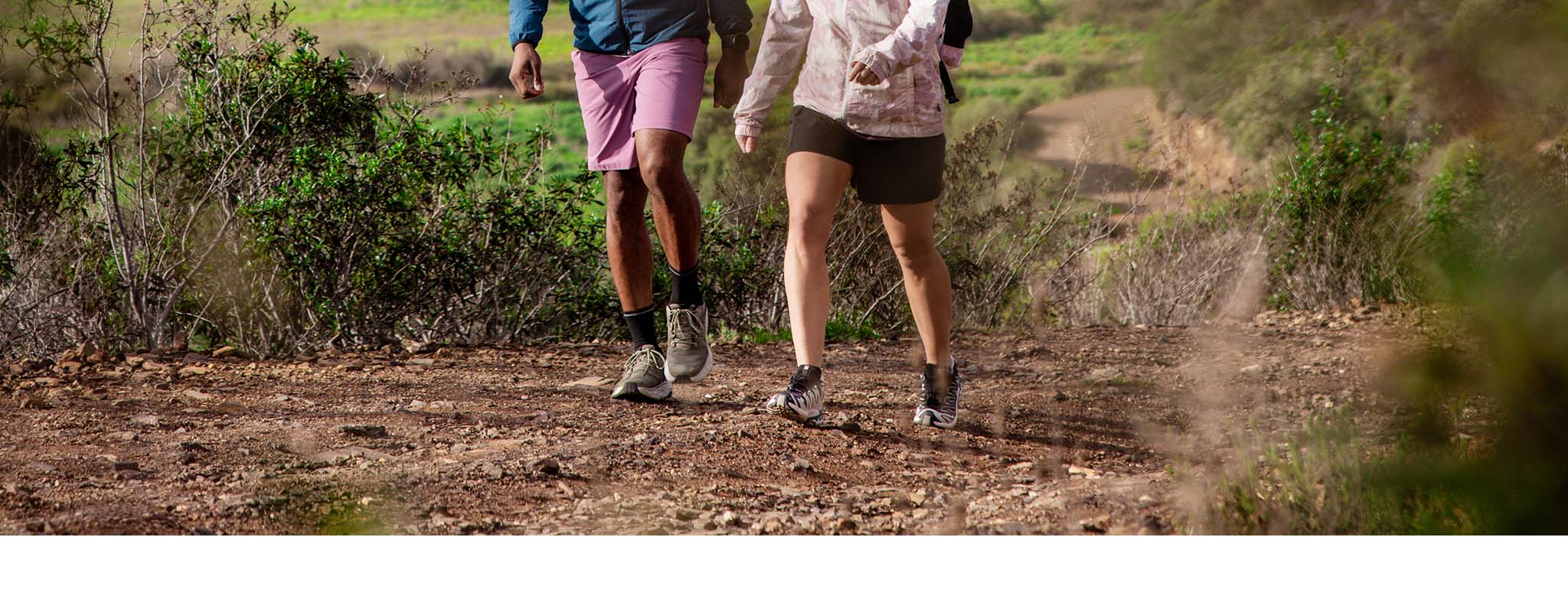 Two walkers walking towards the camera in light coloured walking shoes and shorts in the sun