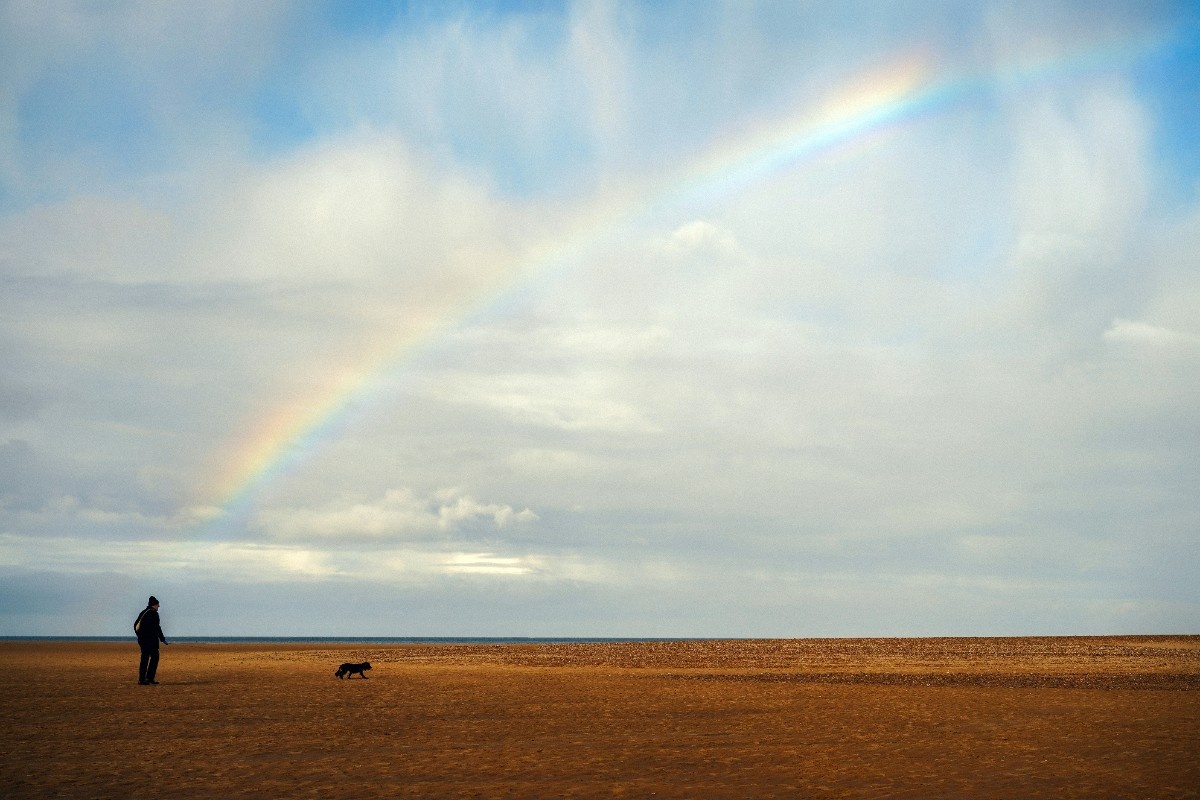 A person walking their dog on Holkham beach with a rainbow in the sky 