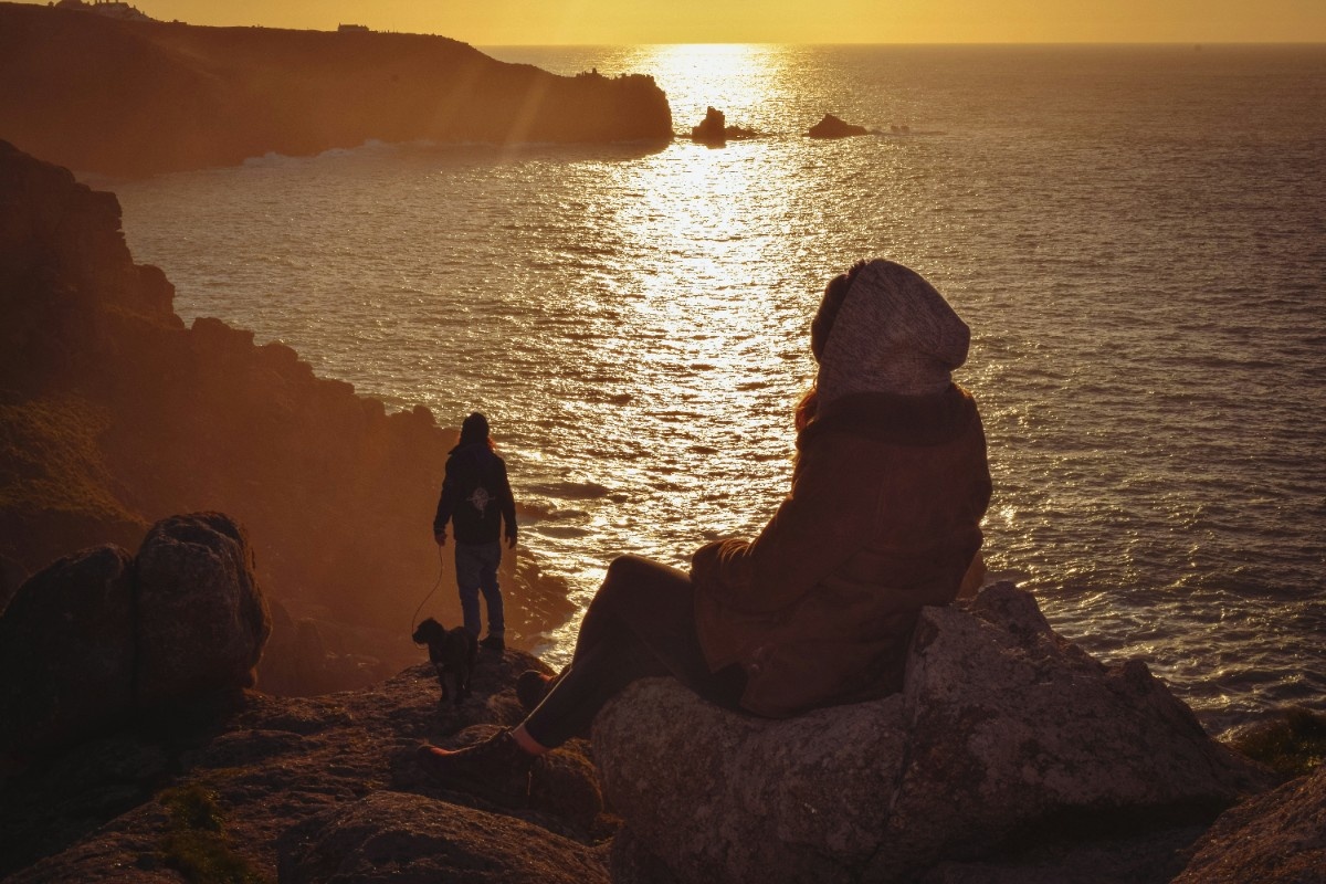 Two people sat on a cliff edge watching the sun set on Sennen Cove