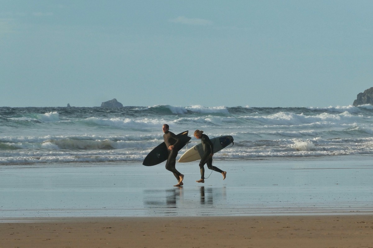 Two people walking along Watergate Bay with their surfboards