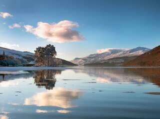 Loch Tay Scotland Crannog on reflective mirror calm fresh water lake in winter with snowy mountains.