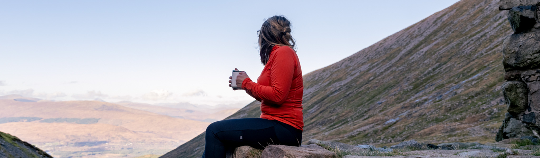 A blonde woman with a bright orange baselayer sits on a rock with a mug looking out over the hills