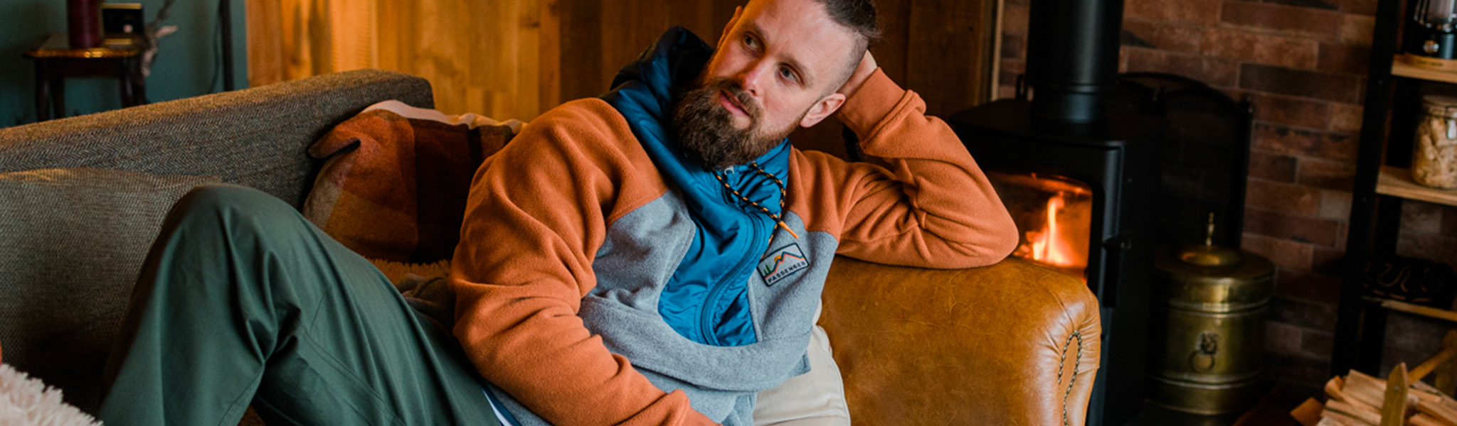 A man in an orange and blue passenger fleece laying on a sofa with a roaring fire behind